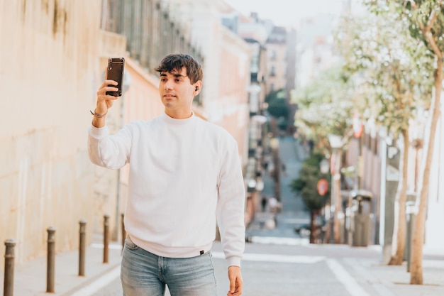 Gai jeune homme espagnol regardant la caméra tenant son téléphone avec des écouteurs dans la rue, portant un pull blanc. Style de vie de liberté de musique de ville. Copiez l'espace avec un arrière-plan flou