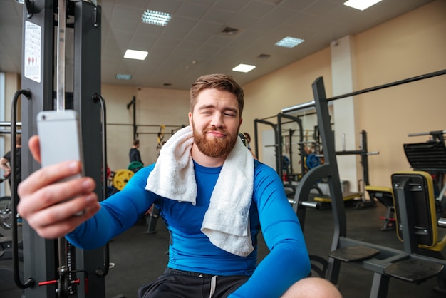 Gai jeune sportif assis dans la salle de gym et faire selfie