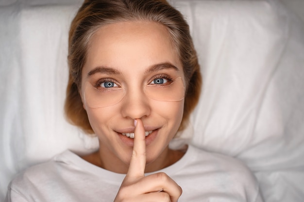 Gaie belle jeune femme allongée sur un canapé blanc. Elle a l'air droite et garde le doigt sur la bouche. Silence. Silence.