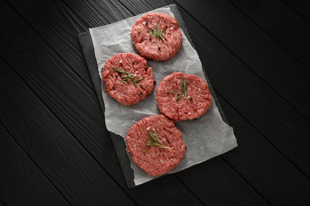 Galettes de burger de boeuf cru sur la planche de bois du boucher romarin et poivre Fond noir Vue de dessus