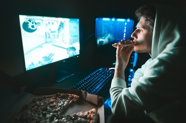 Gamer est assis à la maison à la table à l'ordinateur et mange de la pizza