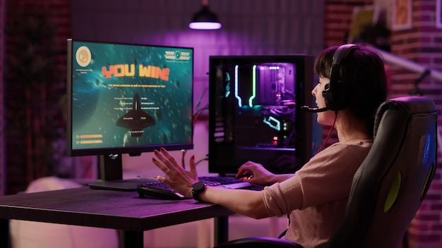 Gamer girl célébrant la victoire de la première place dans le tournoi multijoueur en ligne de tir spatial dans le marié vivant à la maison. Femme jouant à la simulation de jeu d'action sur une configuration pc professionnelle avec clavier de jeu.