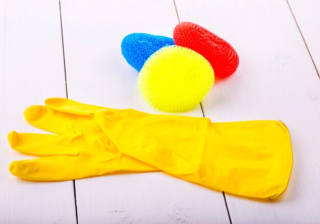 Gants en caoutchouc jaune et éponges colorées sur une table en bois blanche