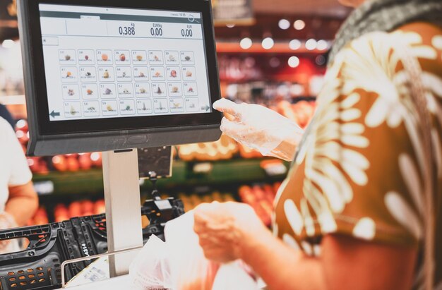 Des gants de protection pour femme aînée pèsent ses achats de légumes sur la balance électronique du supermarché