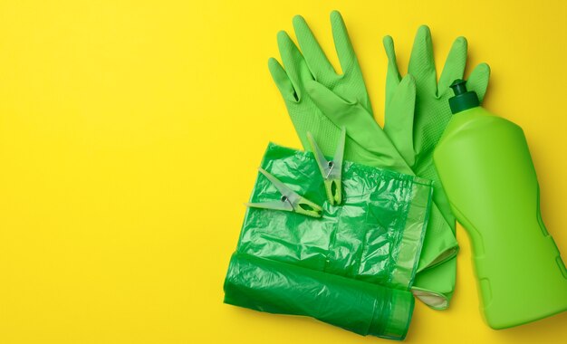 Gants verts en caoutchouc pour le nettoyage, rouleau de sac en plastique poubelle et bouteille en plastique avec détergent sur fond jaune, ensemble