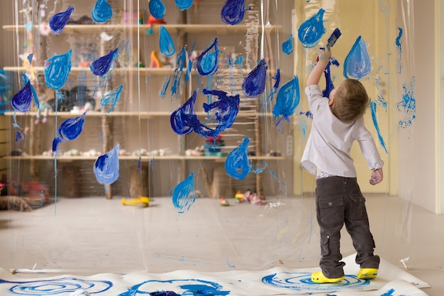 Un garçon de 3 ans en chemise blanche dessine des gouttes de pluie bleues sur un matériau transparent