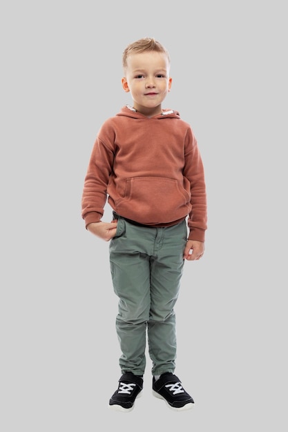 Le garçon a 5 ans. Un enfant en jean et pull marron. Isolé sur un fond gris clair. Pleine hauteur. Verticale.