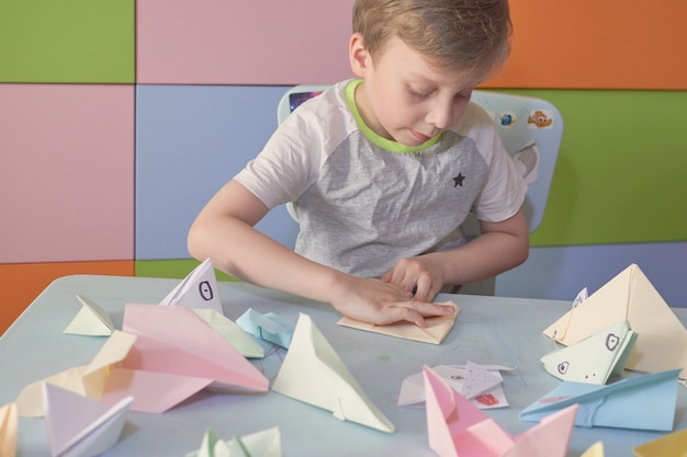 Un garçon de 6 ans fabrique des avions et des grenouilles en origami pendant la quarantaine Covid-19, l'auto-isolement, le concept d'éducation en ligne, l'enseignement à domicile. Enfant à la maison, jardin d'enfants fermé, art pour enfants.