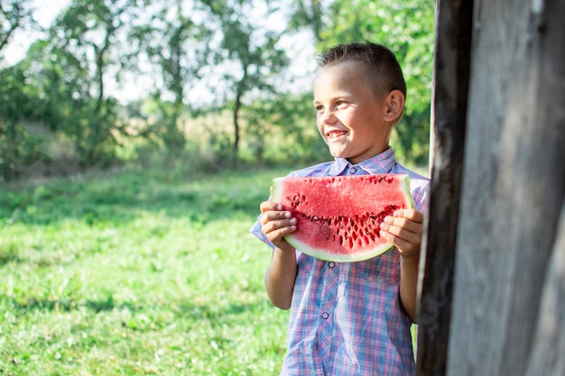 Un garçon de 8 ans se tient dans le jardin du village et tient un gros morceau de pastèque