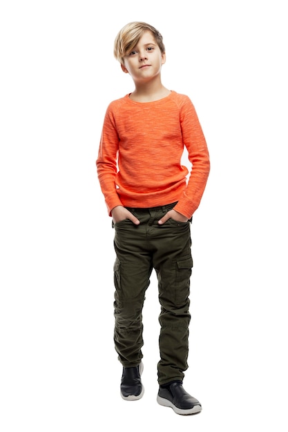 Un garçon de 9 ans vêtu d'un pull orange et d'un pantalon vert se tient les mains dans les poches.