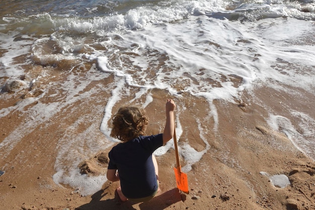 un garçon accroupi devant la mer jouant avec une pelle