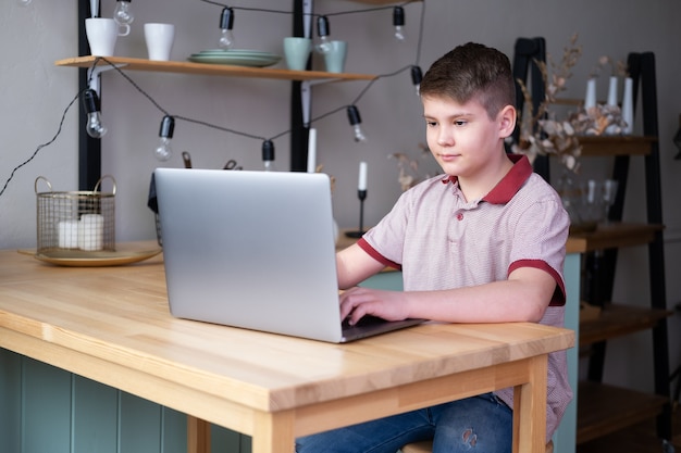 Garçon adolescent étudiant en ligne, jouant à des jeux vidéo à l'aide d'un ordinateur portable assis sur la cuisine.