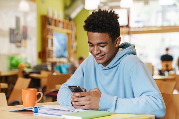 Garçon adolescent souriant étudiant assis au hub à l'intérieur, à l'aide d'un téléphone portable