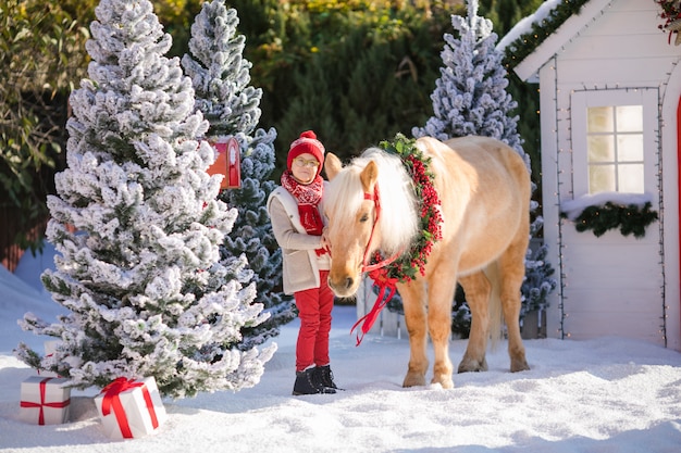 Un garçon adorable avec des lunettes tient son adorable poney avec une couronne de fête près de la petite maison en bois et des arbres enneigés.