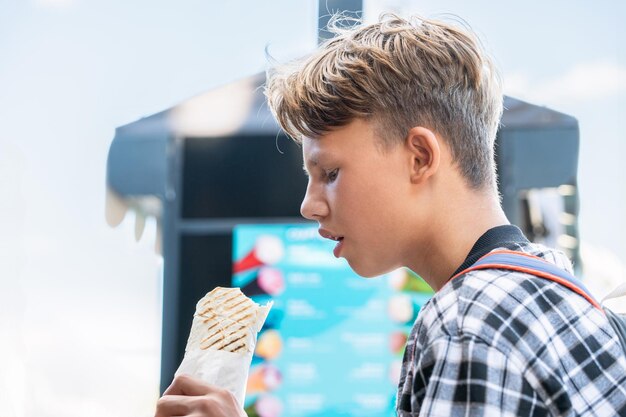 Un garçon affamé dans une chemise à carreaux savourant un savoureux shawarma de poulet dans un parc vert par une journée d'été ensoleillée