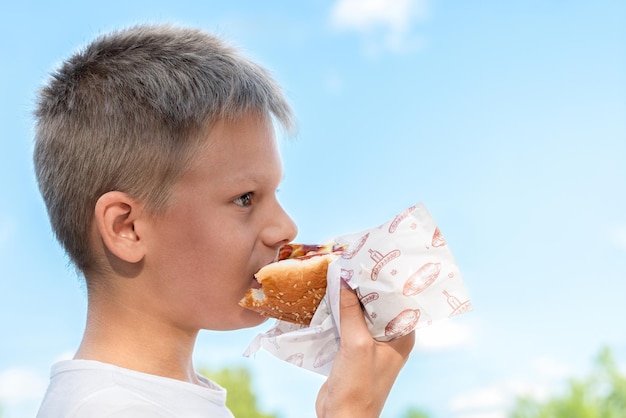 Un garçon affamé savourant un hot-dog à l'extérieur par une journée ensoleillée sur un ciel bleu avec un fond de nuages blancs