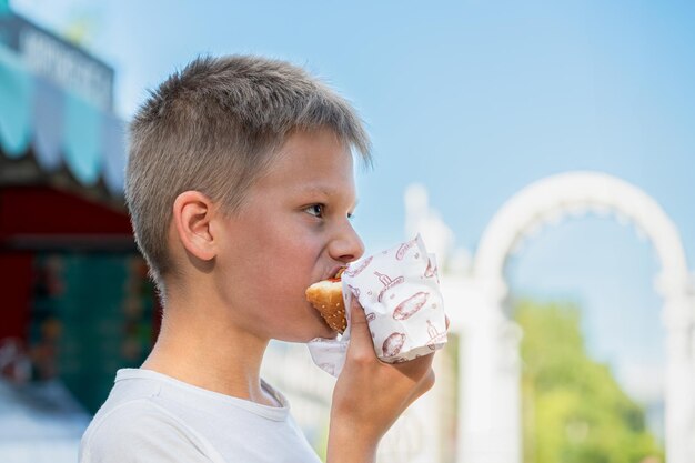 Un garçon affamé savourant un hot-dog à l'extérieur par une journée ensoleillée en prenant une bouchée satisfaisante dans un parc public