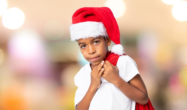 Garçon afro-américain avec chapeau de Noël et prenant un sac avec des cadeaux sur mur flou