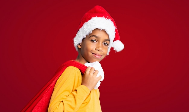 Garçon afro-américain avec chapeau de Noël et prenant un sac avec des cadeaux sur le mur rouge