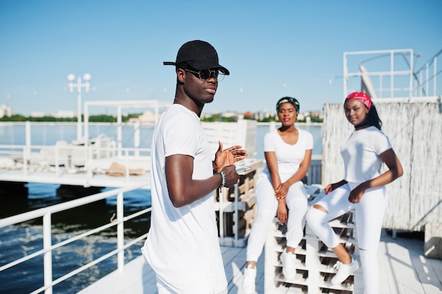 Un garçon afro-américain élégant porte des vêtements blancs, des lunettes et une casquette avec un téléphone portable à la main contre deux filles Mode de rue des jeunes noirs