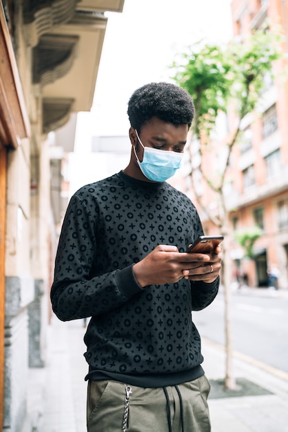 Garçon afro-américain noir marchant dans la rue avec un masque bleu regardant son téléphone portable se protégeant de la pandémie de coronavirus Covid-19