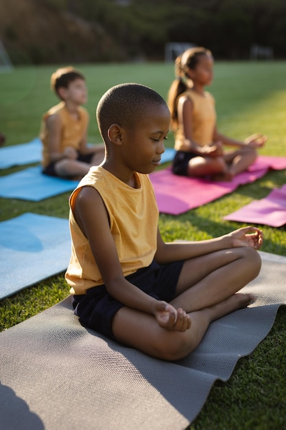 Garçon afro-américain pratiquant le yoga et méditant assis sur un tapis de yoga dans le jardin à l'école