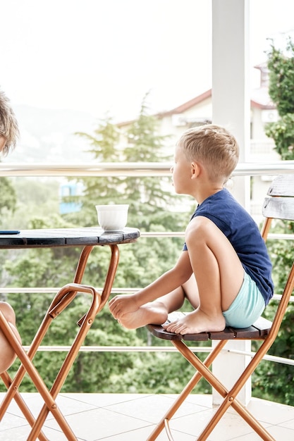 Un garçon d'âge préscolaire boit du lait chaud dans une tasse assis sur une chaise tôt le matin sur le balcon de l'hôtel