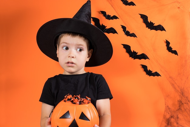 Un garçon d'âge préscolaire en costume de sorcière tient une citrouille avec des bonbons.