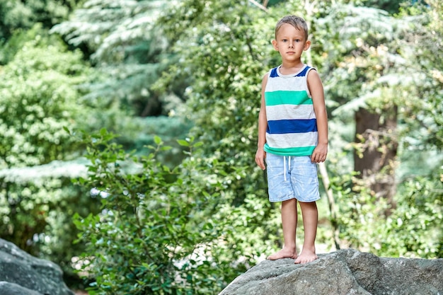 Un garçon d'âge préscolaire ravi se dresse au sommet d'un gros rocher dans le parc de la ville contre des arbres verts flous