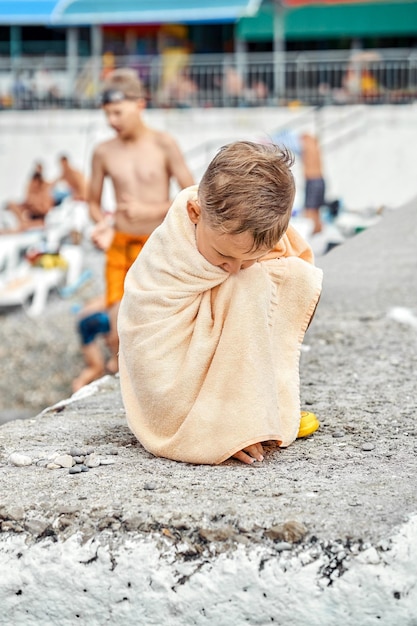 Un garçon d'âge préscolaire recouvert d'une serviette beige s'assoit sur la jetée et se refroidit après avoir nagé en mer