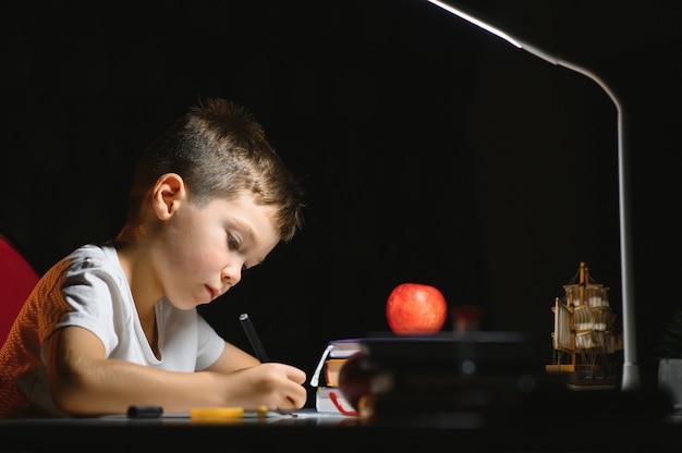 Le garçon apprend des leçons à la maison à la table à la lumière d'une lampe de table.