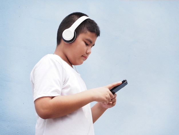 Garçon asiatique avec une chemise blanche portant des écouteurs et jouant du téléphone mobile sur fond de mur bleu.