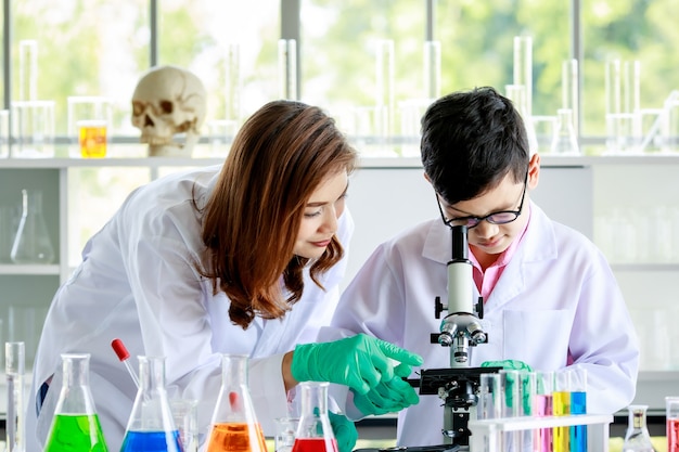 Garçon asiatique concentré examinant des liquides au microscope tout en menant une expérience en laboratoire scolaire avec une enseignante.