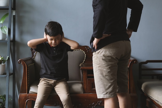 Un garçon asiatique est assis sur une chaise, couvre ses oreilles et ferme les yeux tout en étant grondé par son père