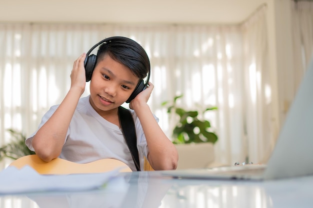 Garçon asiatique jouant de la guitare et regardant un cours en ligne sur un ordinateur portable tout en s'entraînant pour apprendre la musique