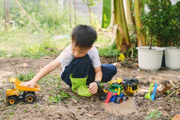 Garçon asiatique portant des bottes jouant dans des flaques boueuses creusant dans un sol boueux avec un camion jouet à la maison