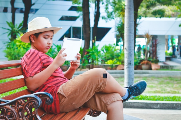 Garçon asiatique portant un chapeau assis sur une chaise jouant une tablette