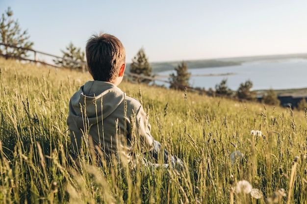 Garçon assis au sommet d'une colline dans un champ d'herbe et profitant d'une belle vue sur le paysage Vue arrière d'un randonneur adolescent se reposant dans la nature Mode de vie actif Concept de voyage local