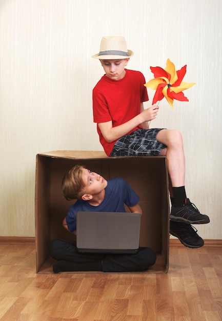 Garçon assis dans une boîte en carton avec un ordinateur portable garçon assis sur le carton avec un papier moulin à vent coloré