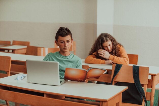 Un garçon assis dans une salle de classe tenant un livre à la main et utilisant un ordinateur portable Mise au point sélective Photo de haute qualité