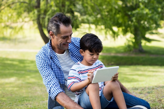 Garçon assis sur les genoux de son père et à l'aide de tablette numérique dans le parc