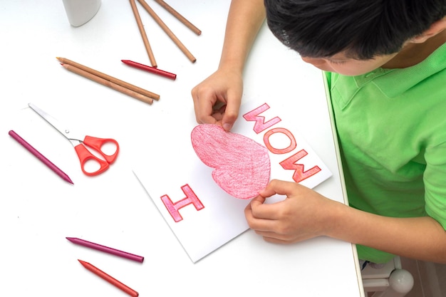 Garçon assis à la table collant un coeur rouge pour le coller sur la carte de fête des mères faite par lui-même