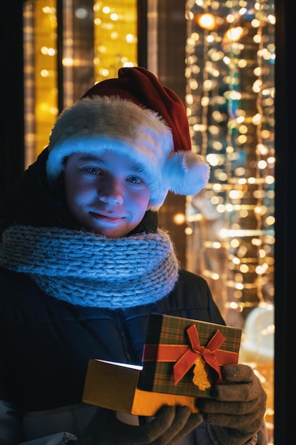 Garçon au chapeau du père Noël avec boîte-cadeaux près de la vitrine illuminée. Noël présente des vacances ou des achats sur le concept du Nouvel An ou de Noël