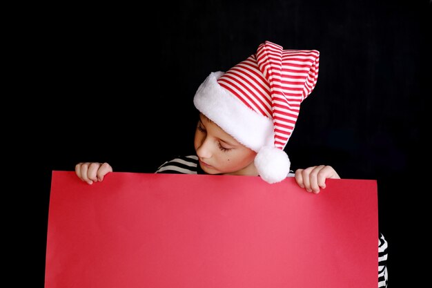 Garçon au chapeau du père Noël tenant le papier blanc rouge pour les voeux de Noël. Isolé sur noir