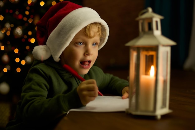 Un garçon au chapeau écrit une lettre au père Noël à la lumière d'une lanterne sur l'arbre de Noël