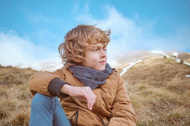 Garçon aux cheveux bouclés dans des vêtements chauds assis sur de l'herbe sèche sous un ciel sans nuages et contemplant le paysage