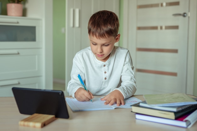 un garçon aux cheveux noirs dans une chemise blanche est assis à une table et fait ses devoirs à l'aide d'une tablette.