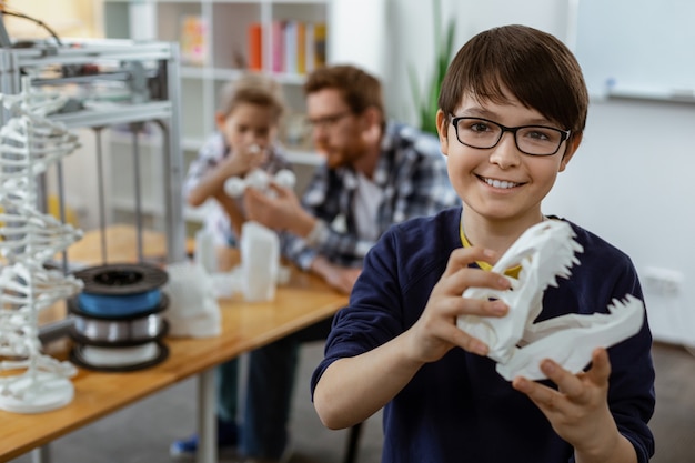 Garçon aux cheveux noirs rayonnant en pull bleu portant un modèle de tête blanc en 3D pendant une leçon innovante