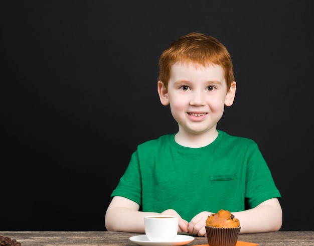 un garçon aux cheveux roux avec un beau visage boit du thé, des grains de café sont allongés à côté de lui, un enfant avec de la nourriture mange un petit gâteau et joue avec du café