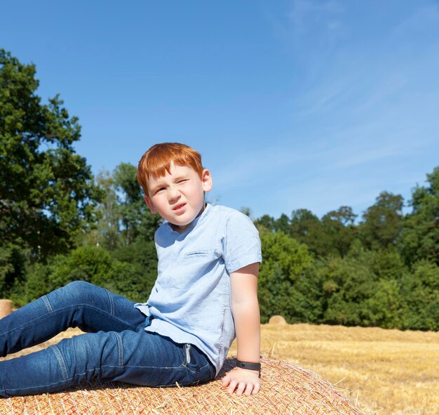 un garçon aux cheveux roux est assis au sommet d'une pile de paille dorée dans un champ, un garçon sur une pile de paille de blé épineux, un portrait d'un garçon de sept ans contre un ciel bleu